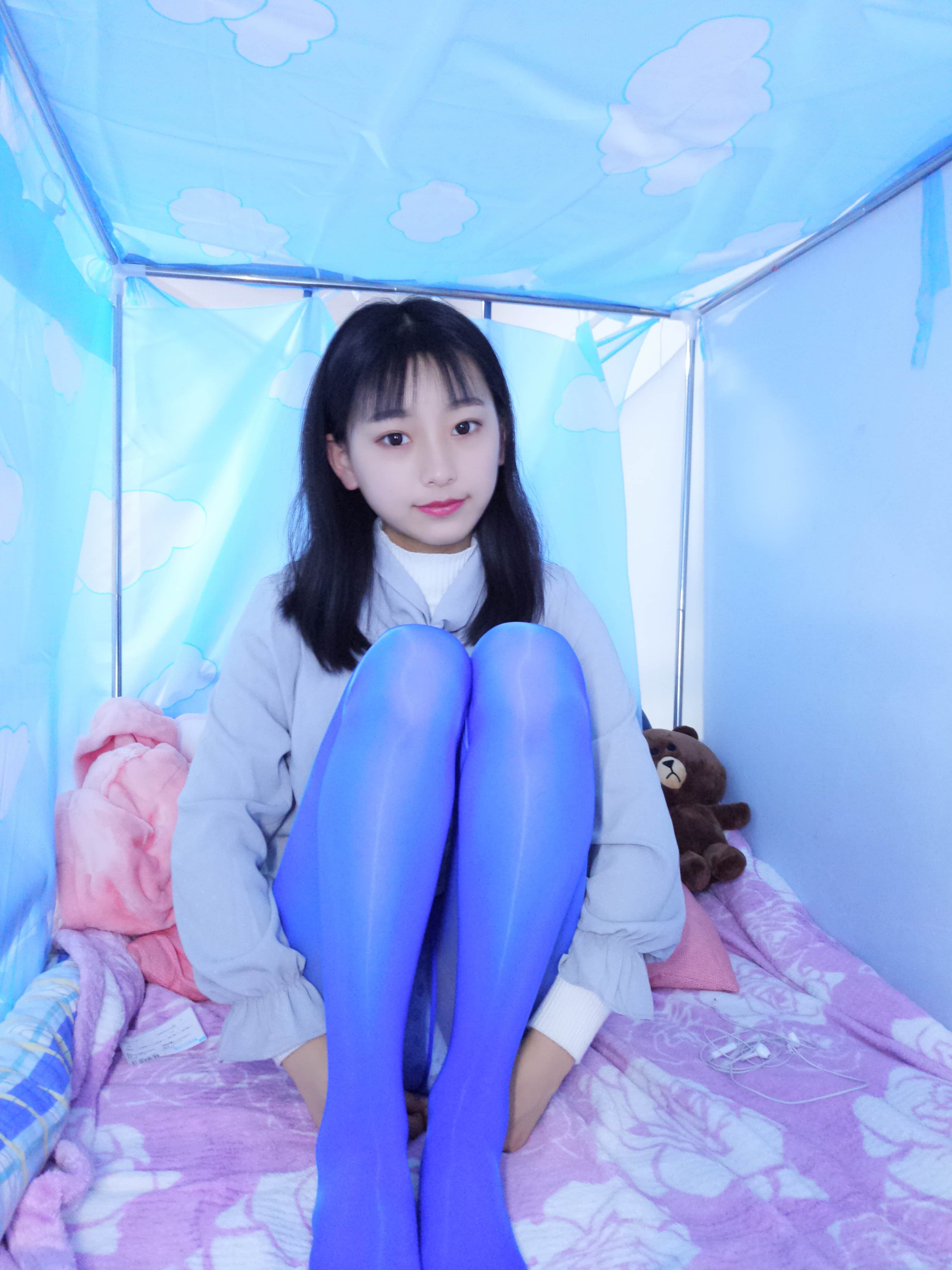 Close-up of glossy blue stockings on arched feet, toes flexing against sheer nylon over soft bedding.