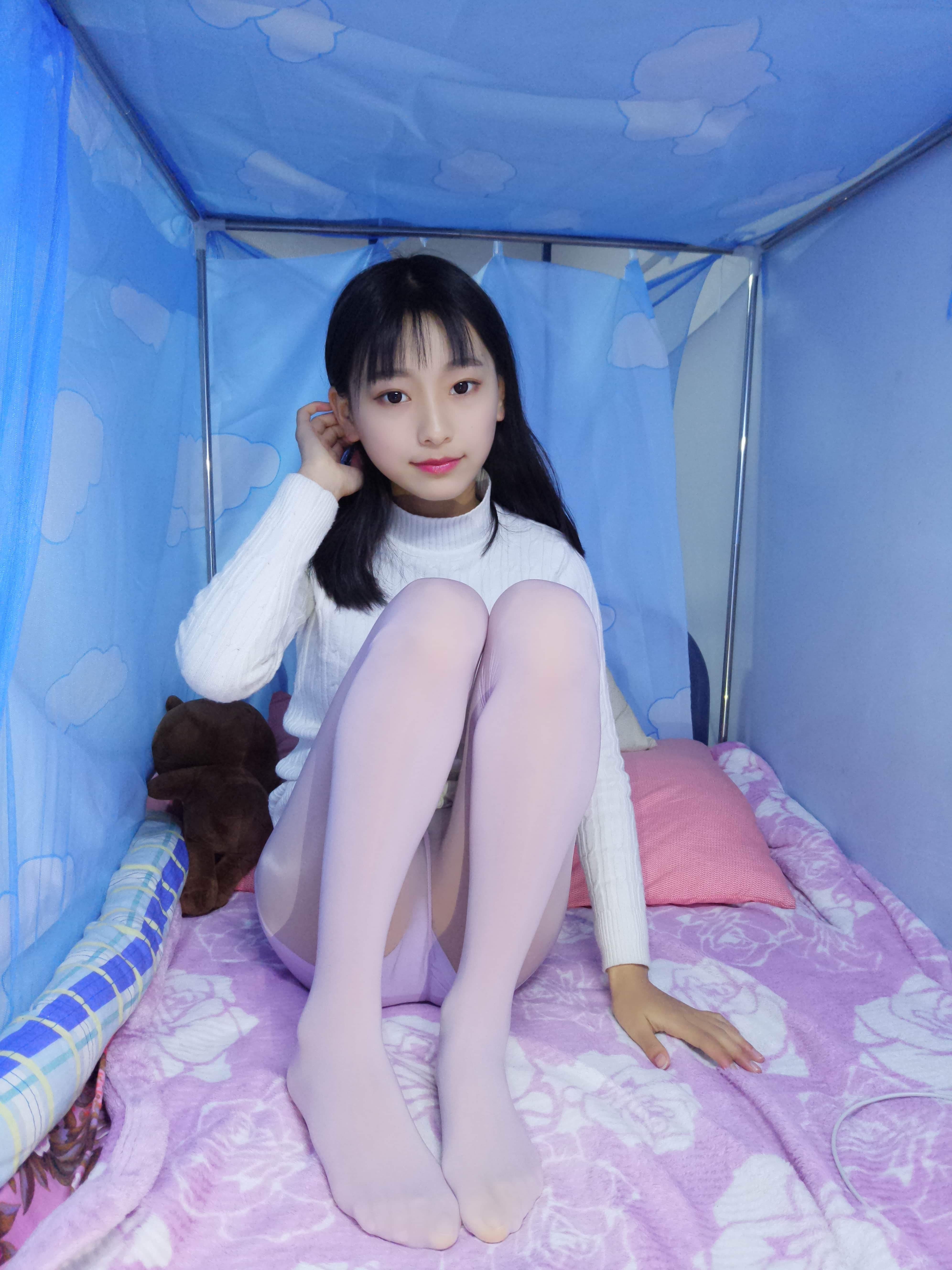 Close-up of beige shiny stockings hugging calves with purple panties peeking, toes resting on soft bedding.