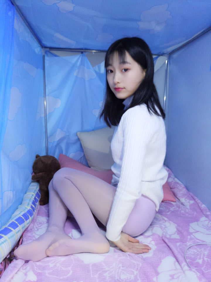 Artful shot of arched feet in beige nylon, purple panties visible, set against a dimly lit rumpled bed.