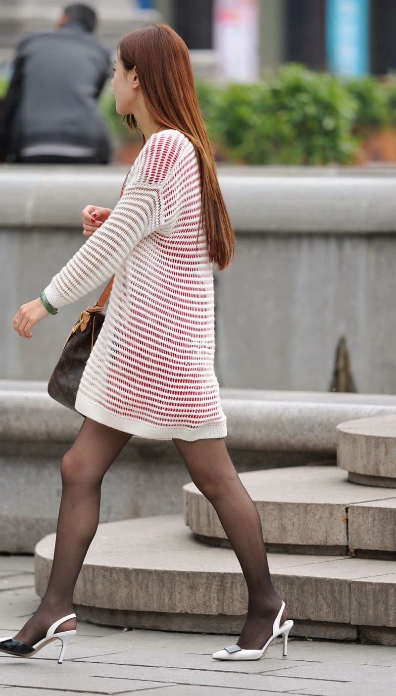 Street portrait of young woman smiling, hands in pockets, black stockings visible beneath short skirt, sunlight and urban blur behind.