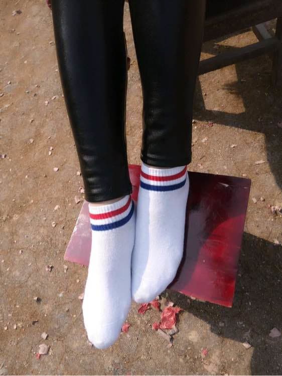 Adult woman outdoors sits on a park bench, peeling off white cotton socks to reveal glossy sheer stockings and bare heels beneath.