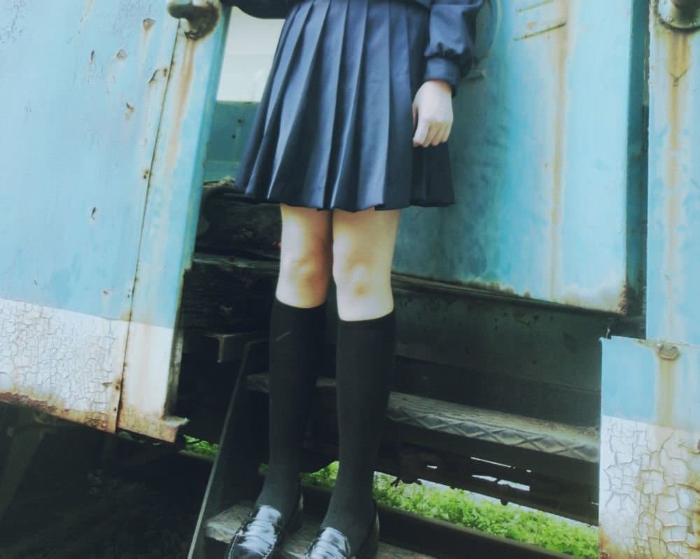 Masked woman seated on a train, sheer stockings visible, one shoe off revealing bare toes and a teasing arch in soft cabin light