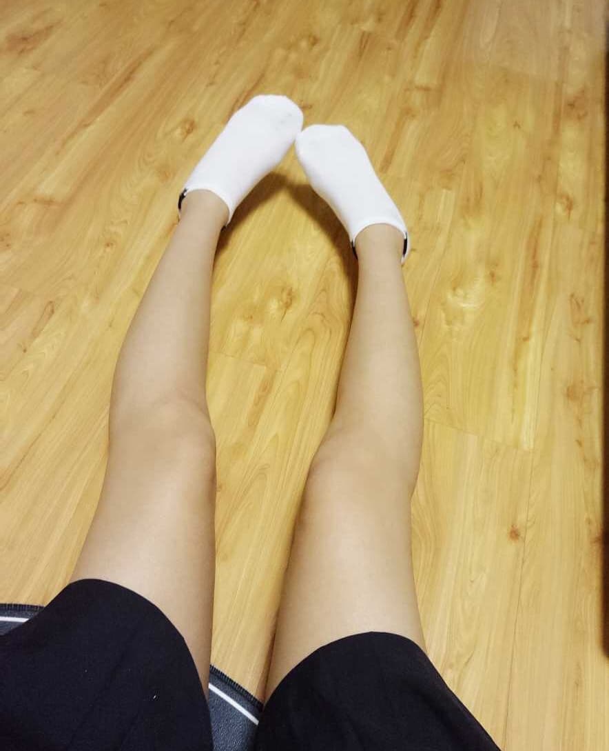 Knee-length pleated skirt with slouched socks and bare feet dangling, soft natural light from a dorm-like window.