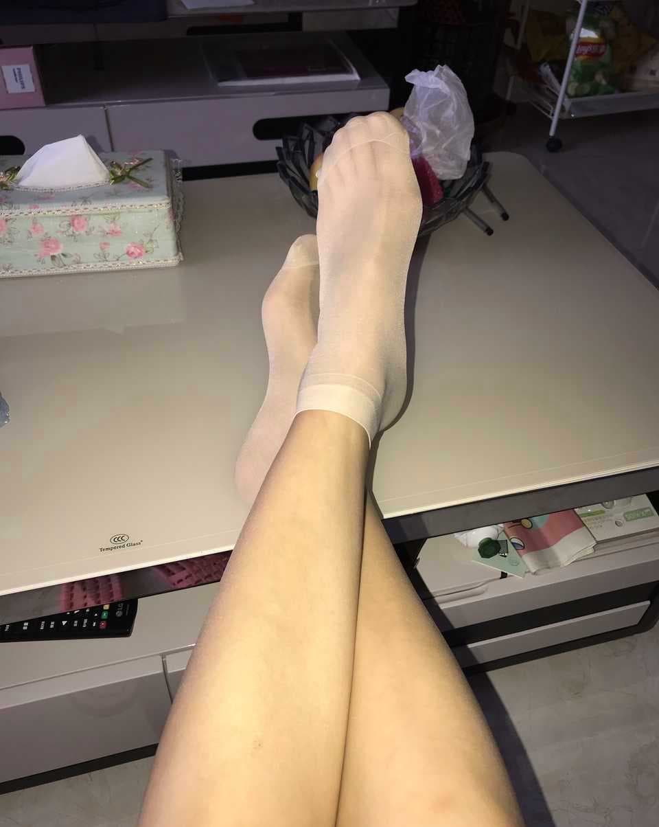 Bare feet lounging on a living room couch, stockings half-rolled, toes relaxed, soft sunlight accentuating smooth skin and nylon sheen.