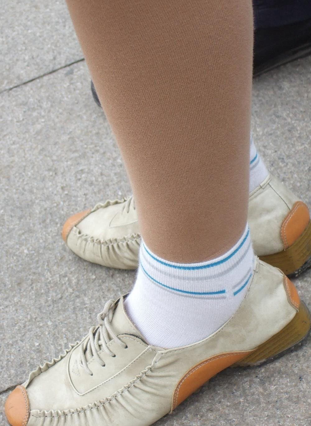 Two women pause on a city sidewalk, one crossing legs to reveal stocking-draped ankle and exposed toes