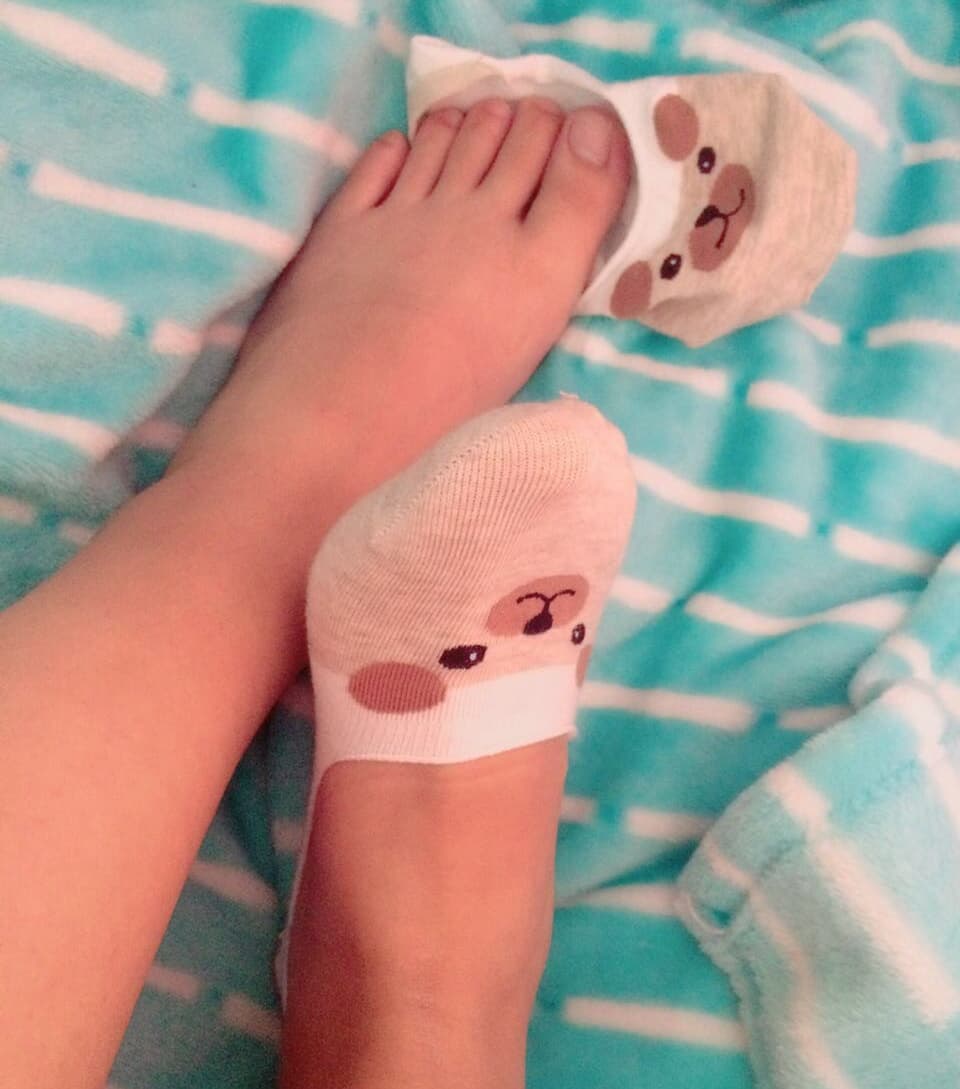 Petite adult figure playfully wiggling toes from under a cozy blanket, bare soles visible in soft light